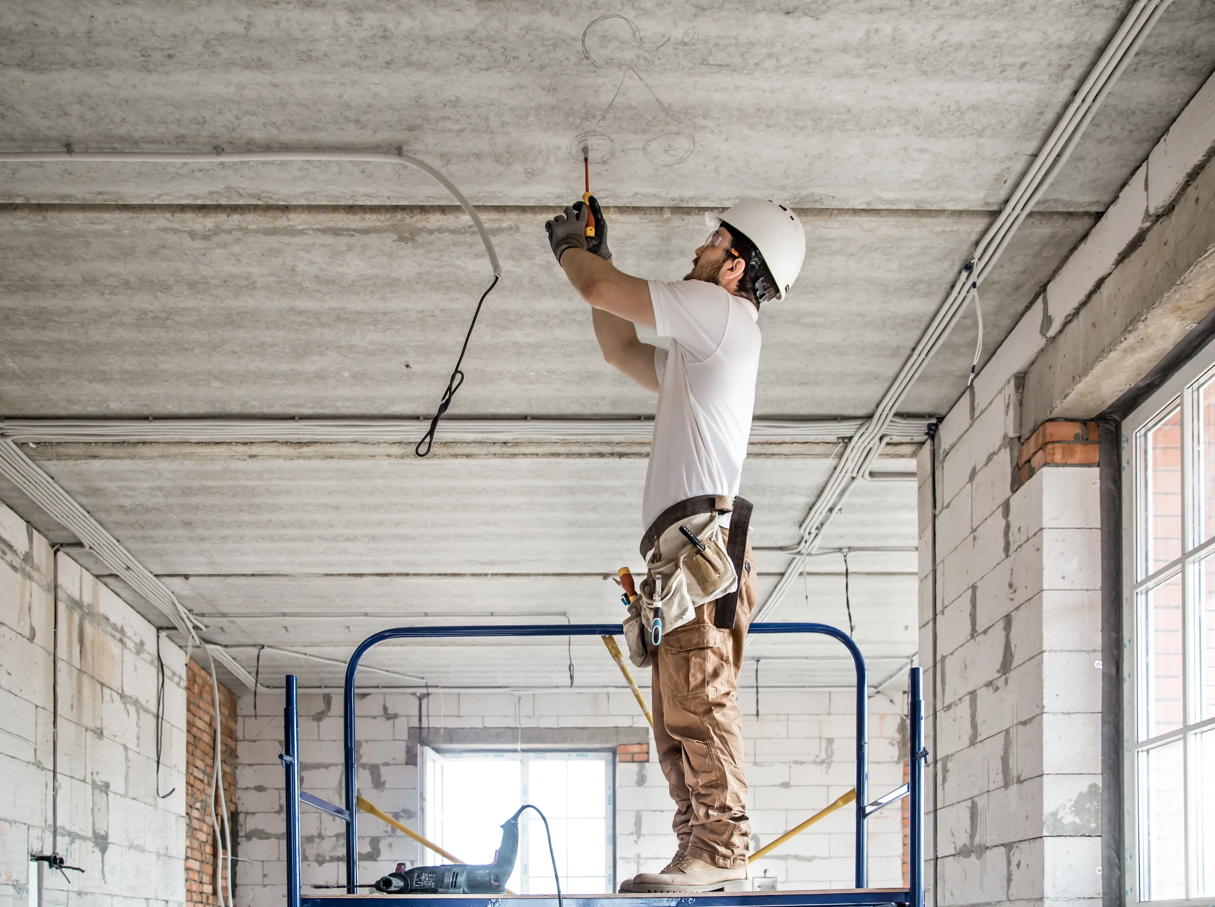 An electrician in a hard hat standing on a mobile work platform, reaching overhead to run cable along a bare concrete ceiling