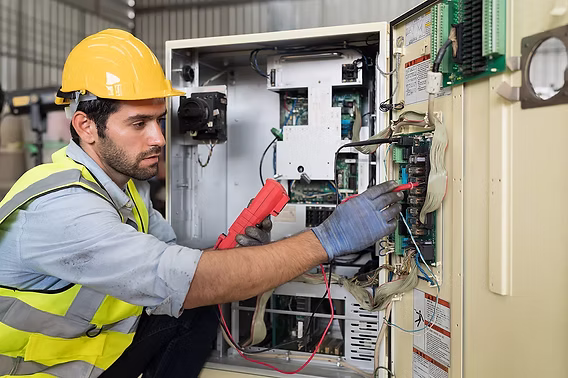 An electrician in a hard hat and high-visibility vest testing an open industrial control panel with a multimeter