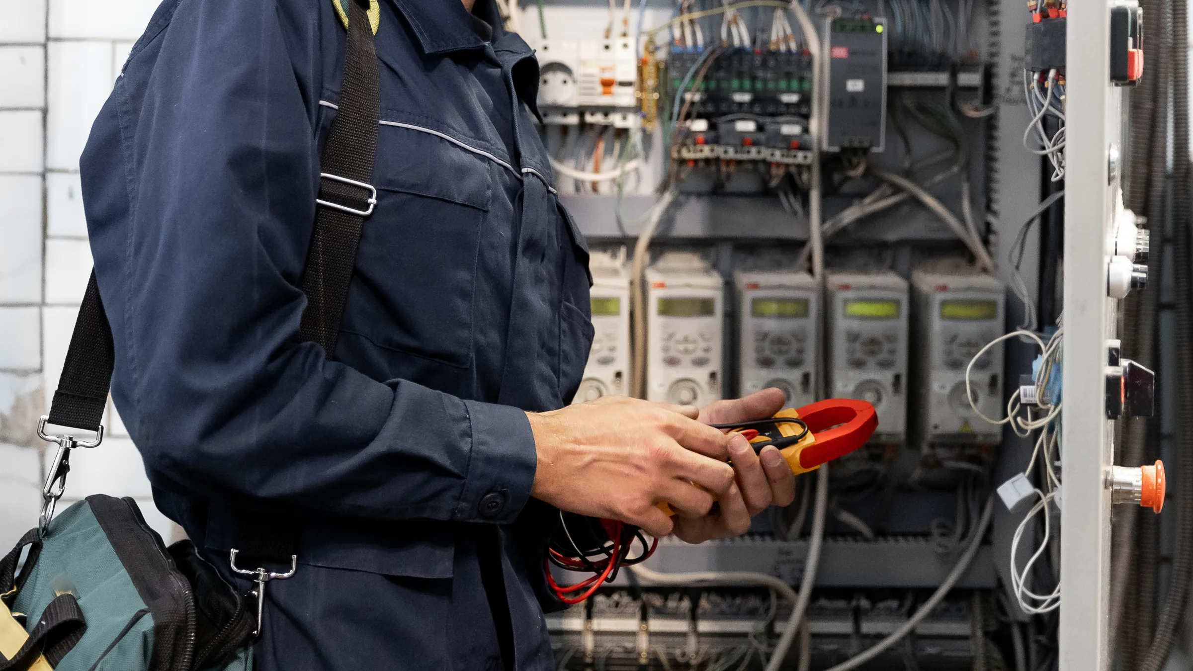 A technician in coveralls holding a clamp meter in front of an open control panel of breakers and wiring