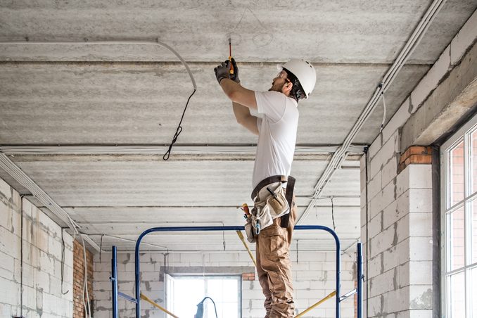 An electrician in a hard hat standing on a mobile work platform, reaching overhead to run cable along a bare concrete ceiling
