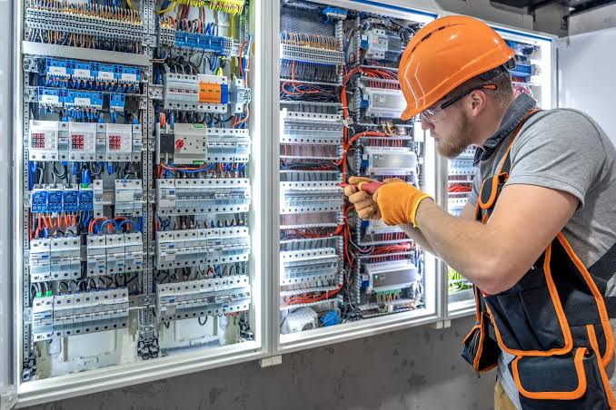 An electrician in a hard hat working inside a large commercial distribution board of DIN-rail breakers and wiring — electrical contractor insurance in Wisconsin