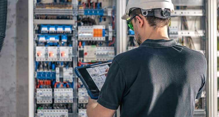 An electrician holding a tablet in front of an open commercial control panel of breakers and wiring — electrical contractor insurance in Nebraska
