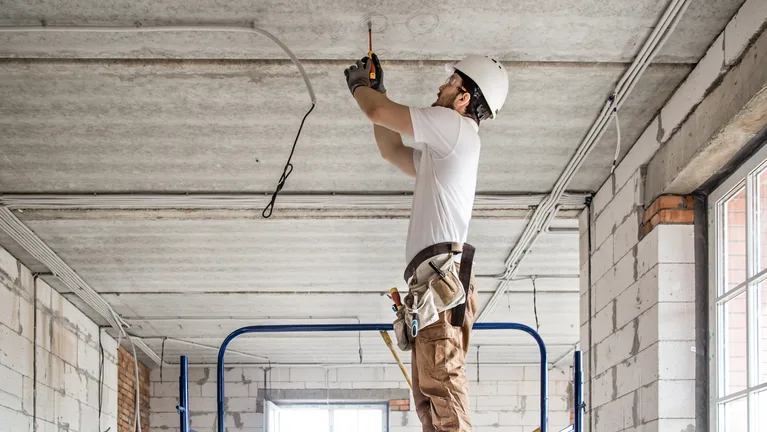 An electrician in a hard hat standing on a mobile work platform, reaching overhead to run cable along a bare concrete ceiling — electrical contractor insurance in New York
