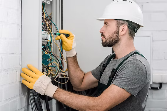 An electrician in a hard hat and gloves working on an open wall-mounted junction box with a screwdriver — electrical contractor insurance in Iowa